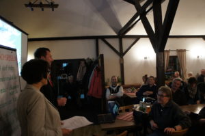 Planer vor Flipchart und Beamerwand, rechtsseitig Ausschnitt der Veranstaltungsteilnehmer